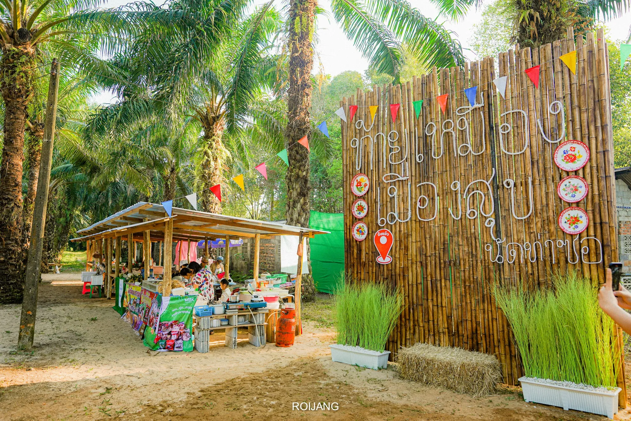 ป้ายไม้ไผ่แสดงคำขวัญ “ท้ายเหมือง เมืองสุข หลาดนัดท่าซอ” พร้อมบรรยากาศแผงร้านอาหาร และร้านขายสินค้าพื้นเมือง